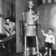 Child holds hands with robotic "mom" while a man operates controls in the background.