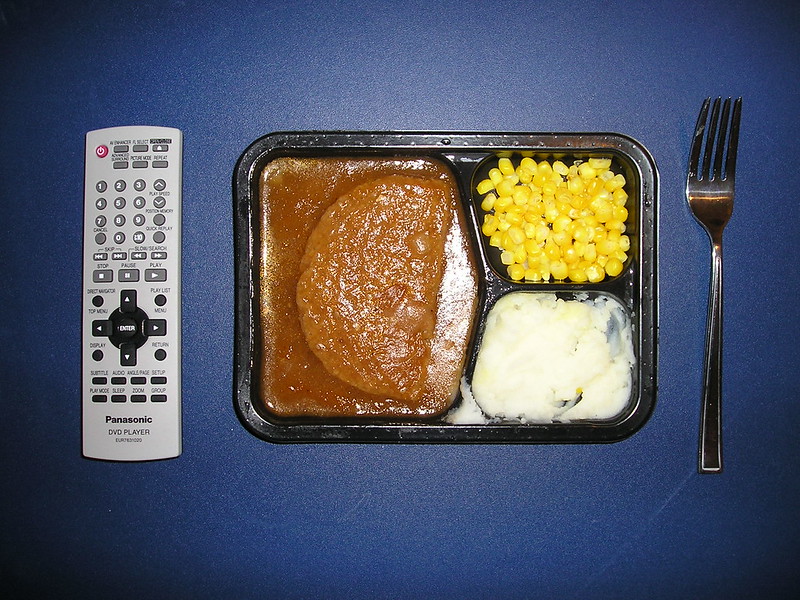A TV dinner (meatloaf in gravy, corn, and mashed potatoes) in a black plastic tray, with a remote control to the left and silverware to the right. (Photo by John Atomic via Flickr/Creative Commons https://flic.kr/p/jKJNF)