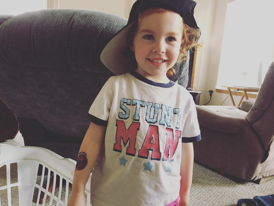 Two year old is wearing her hat sideways and smiling. Also she is wearing a shirt that says "STUNT MAN"