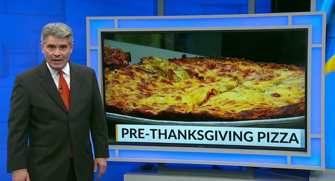 News anchor in front of an image of a pizza with the caption "Pre-Thanksgiving Pizza"