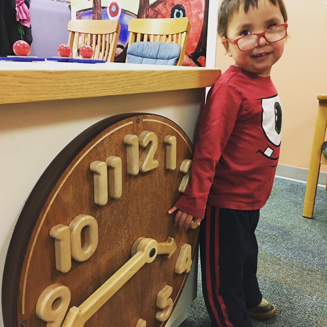 Almost three year old stands next to a big toy wooden clock with a big smile on his face.