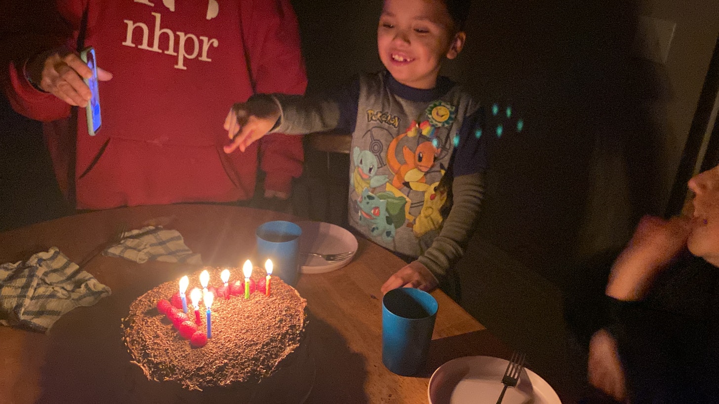 Seven year old smiles and points at his birthday cake, which is chocolate and raspberry and has the number 7 laid out in raspberries and birthday candles.