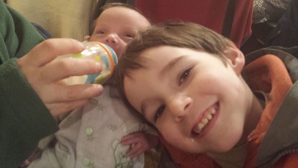Baby boy is drinking a bottle, while big bro is smiling next to him.