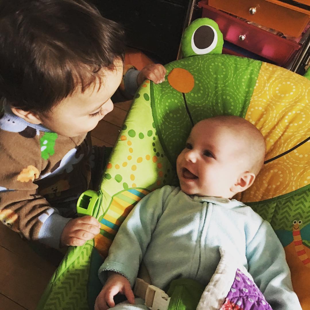 One year old looks at his baby sister in her froggy chair and they are both smiling big smiles at each other.