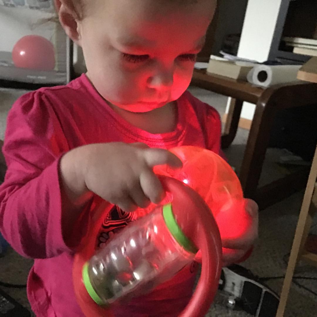 Baby girl looks intently at a glowing red ball while holding a red rattle.