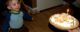 One year old is smiling as he looks at his birthday pie, which has banana pieces on top of whipped cream, plus a birthday candle.