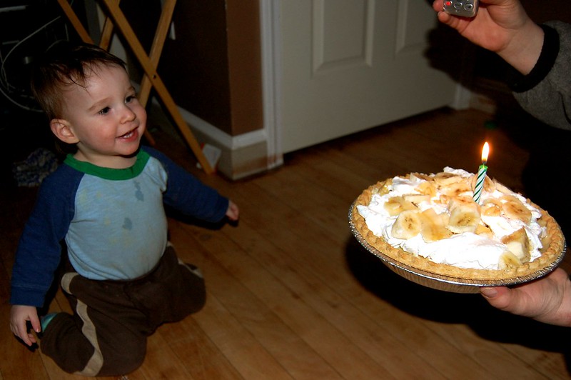 One year old is smiling as he looks at his birthday pie, which has banana pieces on top of whipped cream, plus a birthday candle.
