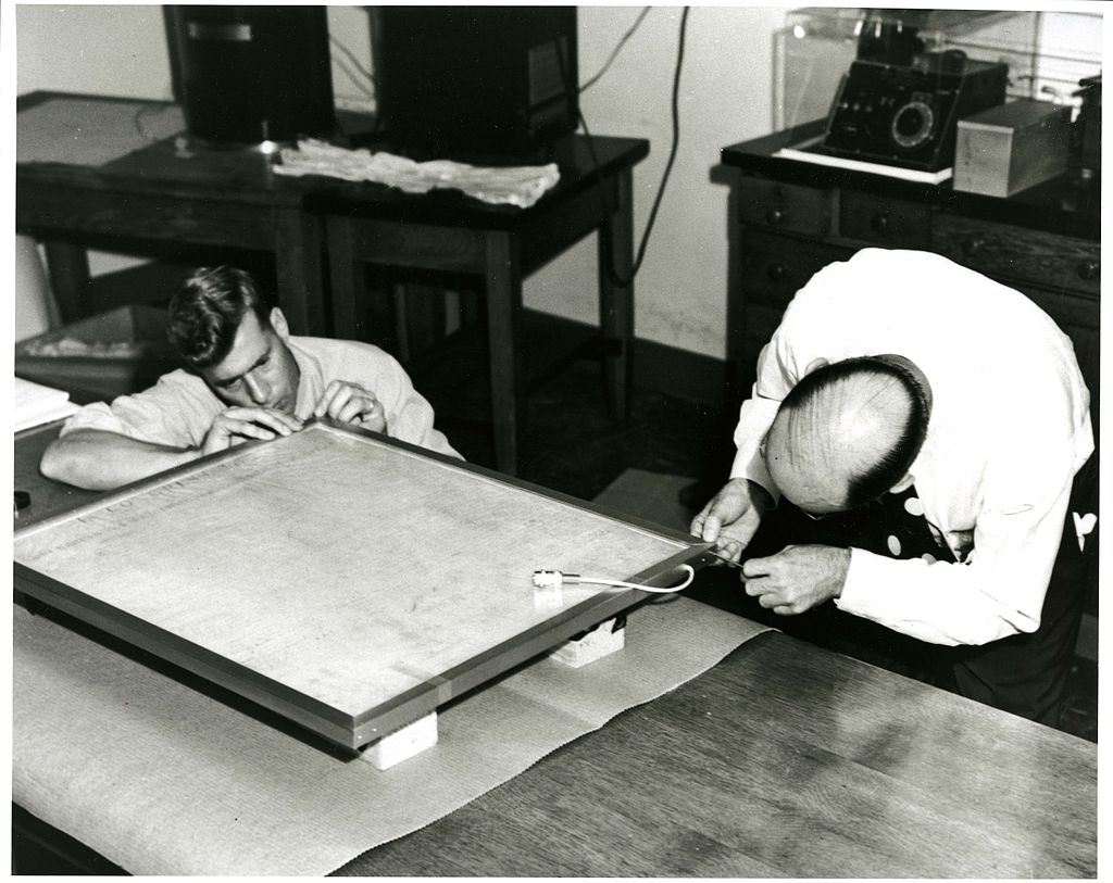 The outer gold frame was placed over the completely enclosed and sealed Declaration of Independence after the flushing operation was completed. Mr. Gilles and Dr. Wampler of Libbey-Owens-Ford Glass Company fasten the last screws in the assembly. The lead from the bottom of the container enabled the leak detector meter to be connected to the leak detecting cells without removing the enclosure from the Shrine at the Library of Congress. Photo via Wikicommons https://commons.wikimedia.org/wiki/File:ChartersOfFreedom_024.jpg