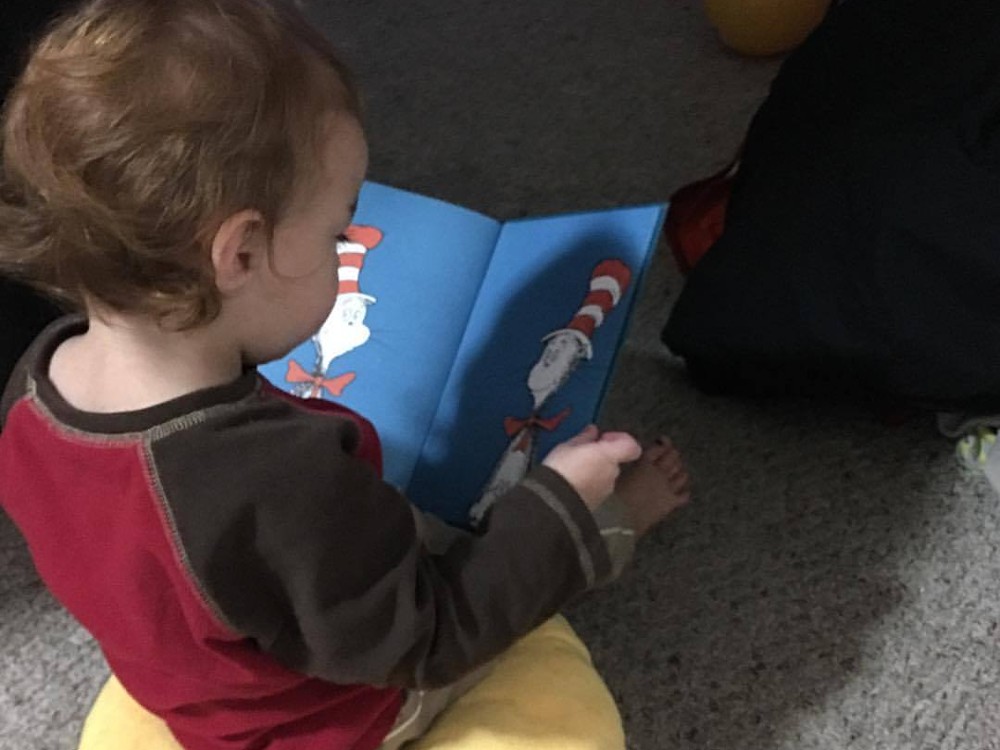 One year old is sitting on her yellow emoji pillow and reading a Cat In The Hat book.