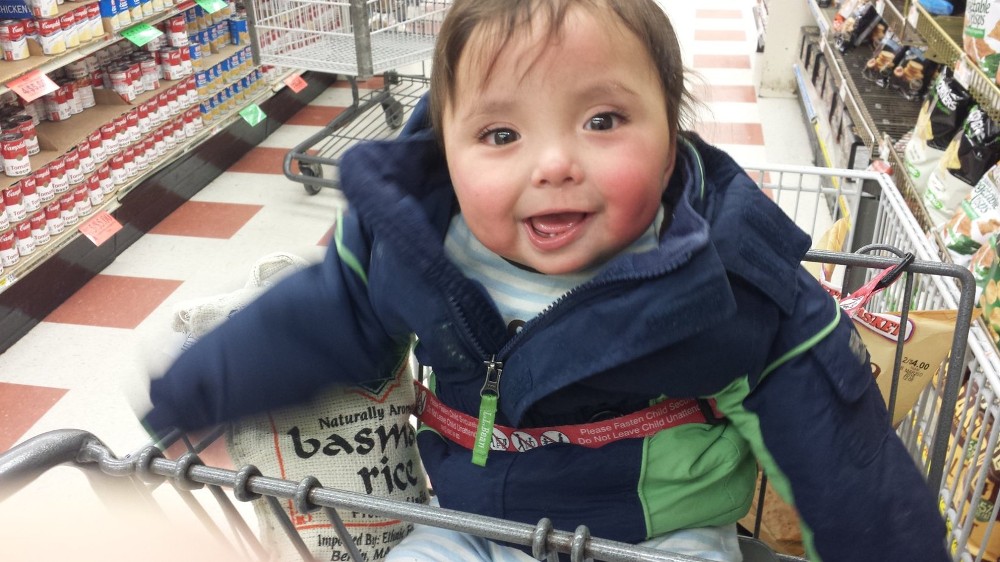 Baby boy is smiling in a shopping cart. Under his right arm is a large bag of basmati rice.