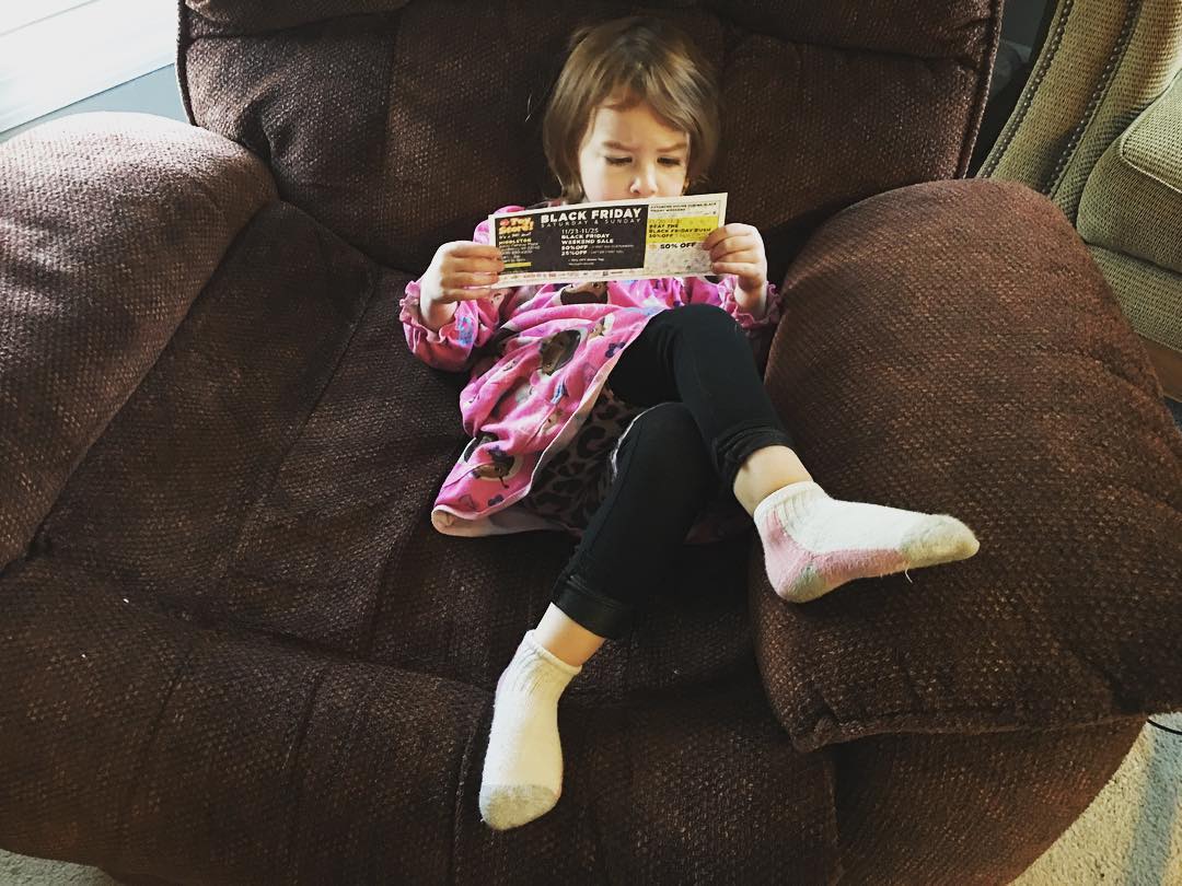 Two year old sits in a big brown recliner, very seriously studying a coupon.