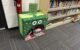 A cardboard box is decorated green with big cartoon eyes, colorful polka dots and a large semicircular "mouth" that holds piles of library books. The sign on top reads "Please give unwanted books to the book monster"